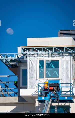 Lavoratore di costruzione in un ascensore blu che lavora all'esterno di un nuovo edificio che installa finestre con un cielo blu e la luna visibile Foto Stock