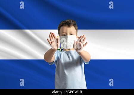 Piccolo ragazzo bianco in una maschera protettiva sullo sfondo della bandiera di El Salvador. Fa un segno di stop con le mani, stare a casa El Salvador. Foto Stock