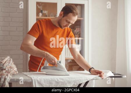 Giovane uomo caucasico che stira le lenzuola dei bambini sull'asse da stiro a casa. Uomini che fanno i chores domestici Foto Stock