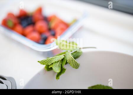 Foglie di menta sul bordo della ciotola bianca con bacche sfocate in un contenitore di plastica come sfondo. Fragole, mirtilli e lamponi in contenitori Foto Stock