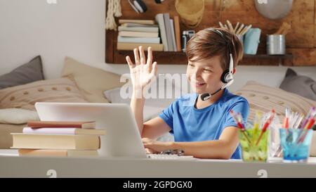 sorridendo ragazzo studentesco che studia a casa con una scuola remota connessa al computer sul web, alza la mano per rispondere alle domande o salutare i suoi compagni di classe, Foto Stock