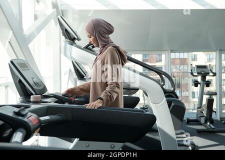 Vista laterale shot di bella donna musulmana che indossa hijab passare il tempo in palestra in piedi sul tapis roulant regolazione impostazioni Foto Stock