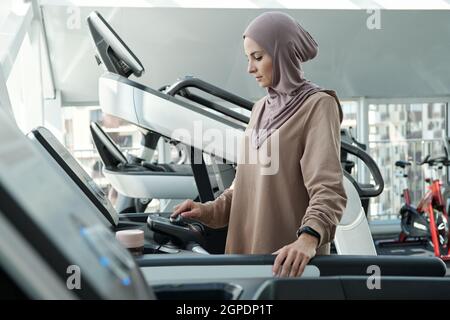 Vista laterale shot di moderna donna musulmana che indossa hijab a partire mattina con l'esercizio in palestra in piedi sul tapis roulant regolazione impostazioni Foto Stock