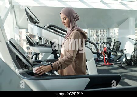 Vista laterale medio shot di giovane adulto musulmana donna che indossa Headwrap iniziare esercizio in palestra in piedi sul tapis roulant regolazione impostazioni prima di correre Foto Stock