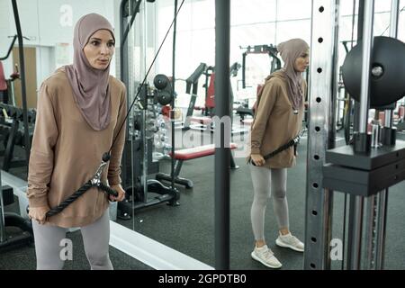 Ritratto di bella donna musulmana sportiva che indossa hijab facendo esercizio di rafforzamento con cavi e pulegge in palestra Foto Stock