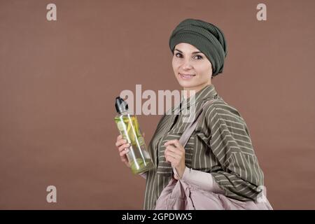 Orizzontale medio ritratto studio di giovane adulto elegante musulmana donna che tiene una bottiglia di limonata fatta in casa fresca sorridendo alla macchina fotografica, sfondo marrone Foto Stock