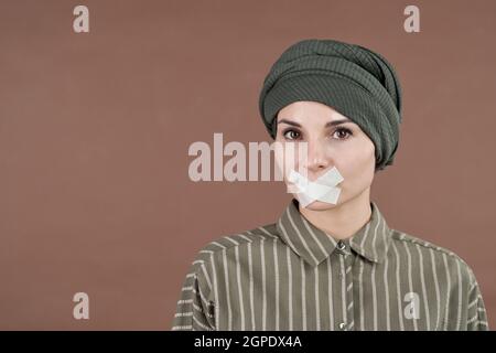 Ritratto concettuale orizzontale medio primo piano di una donna musulmana che indossa il turbante in piedi con nastro sulla bocca guardando la macchina fotografica Foto Stock