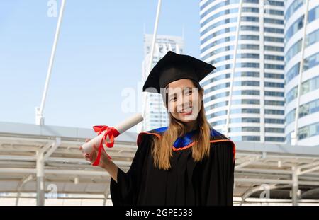 Giovane donna in abito di laurea e cappello con sorriso di felicità e certificato accademico in un giorno di inizio. Concetto di graduazione Foto Stock