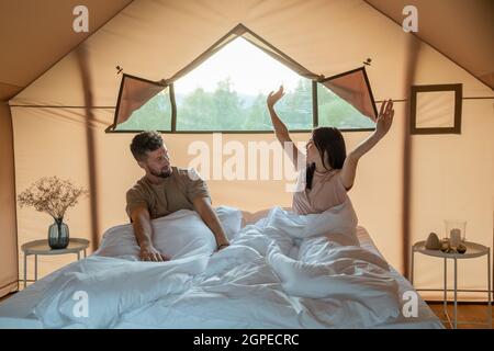 Felice giovane coppia che allunga le braccia dopo il risveglio mentre si siede a letto all'interno di una tenda glamping Foto Stock