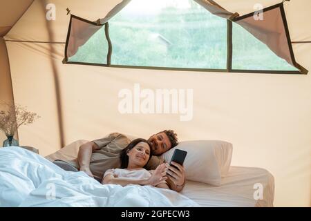 Coppia felice che fa selfie o guarda video sullo smartphone mentre si rilassa a letto Foto Stock