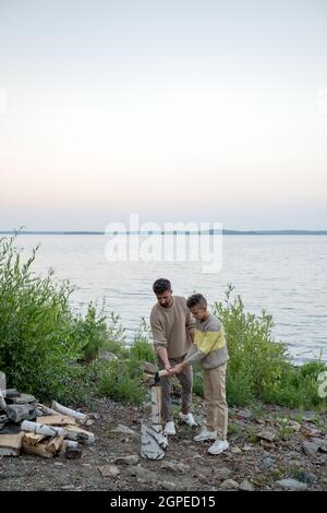 Ragazzo adolescente aiutando suo padre a tritare legna mentre entrambi tenendo l'ascia su log Foto Stock