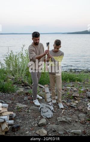 Padre e figlio con ascia tagliando legna insieme contro lago e cielo Foto Stock