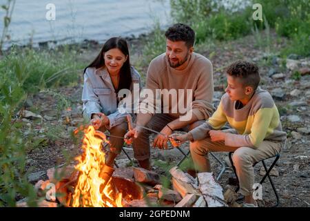 Padre, madre e figlio che tengono spiedini con salsicce sul fuoco mentre cucinano uno spuntino Foto Stock