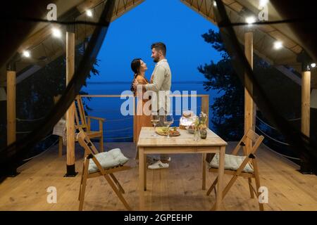 Giovane coppia in piedi in abbraccio sulla terrazza di casa glamping di fronte al tavolo servito Foto Stock