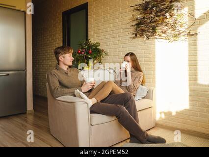 Bella coppia in maglioni bere una tazza di caffè sul divano. Decorazione di Natale sullo sfondo, rami di abete. Donna e uomo che parlano e sorridono. Foto Stock