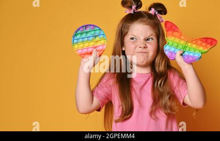 Ragazza di capretto grimacing frolic in camicia rosa tiene due giocattoli sensoriali del colore dell'arcobaleno - schioccalo e guarda da parte al tondo uno Foto Stock