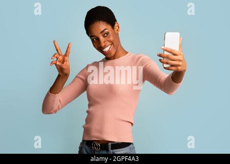 Giovane ragazza allegra che prende Selfie su smartphone e mostra la Pace Gesture Foto Stock