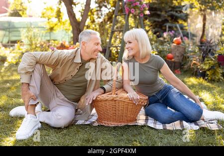 Bella coppia anziana che ha pic-nic all'aperto, seduta su coperta e sorridente l'uno con l'altro, godendo la calda serata autunnale Foto Stock