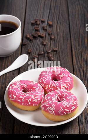 Tre ciambelle rosa con spruzzi in piatto e una tazza di caffè su un vecchio tavolo di legno Foto Stock