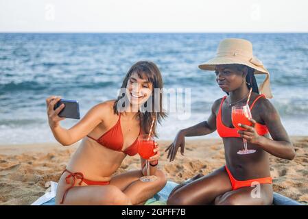 Allegro giovane donna multirazziale migliori amici in bikini rosso prendendo selfie su smartphone mentre rilassarsi con bicchieri di bevande sulla spiaggia sabbiosa vicino al mare durante le vacanze estive Foto Stock