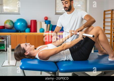 Donna stesa sul letto, facendo esercizi con barra medica per rafforzare i gomiti e le spalle con l'aiuto di un fisioterapista maschile Foto Stock