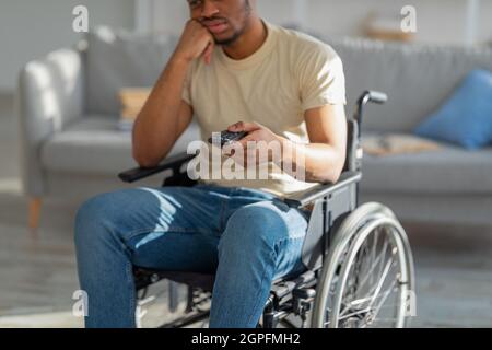 Annoiato uomo nero disabili in sedia a rotelle guardare film noioso in TV, cambiare canale, stanco di televisione a casa Foto Stock