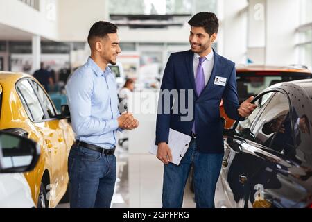 Ragazzo medio-orientale che sceglie la nuova auto, avendo conversazione con il responsabile delle vendite Foto Stock