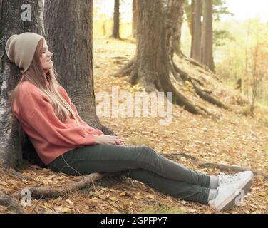 felice giovane donna dai capelli rossi sorridente e seduta sotto l'albero. camminando in una foresta autunnale. viaggio locale. trascorrere il tempo in un parco, rilassante e havi Foto Stock