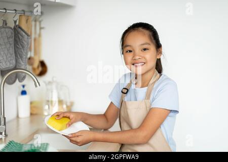 Piccola governante. Ragazza asiatica felice aiutare la madre in cucina, lavando e strofinando piatti, in piedi in cucina Foto Stock