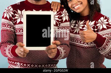 Concetto di vendite di Natale. Uomo nero che tiene il tablet con schermo vuoto e donna che mostra il pollice su sfondo blu Foto Stock