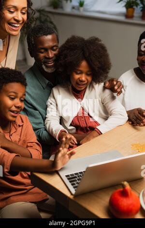 ragazzo afroamericano che agitava la mano vicino al portatile e sorride la famiglia durante la videochiamata il giorno del ringraziamento Foto Stock