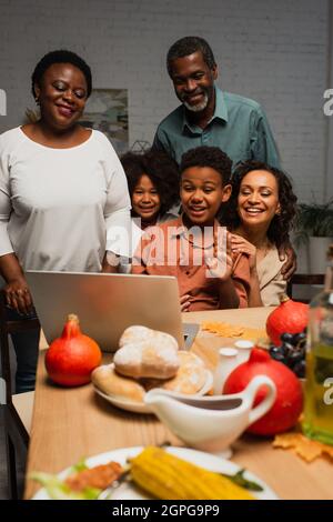 allegro ragazzo afroamericano che agita mano durante la videochiamata vicino alla famiglia e ha servito la cena Foto Stock