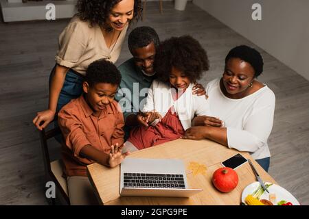 felice ragazzo afroamericano che agitava la mano vicino alla famiglia durante la videochiamata sul laptop Foto Stock