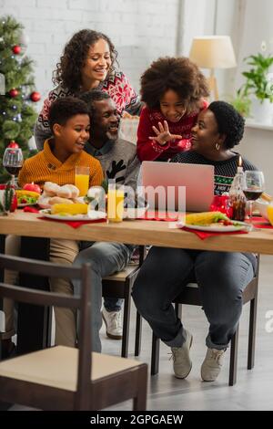 ragazza afroamericana che agita mano vicino al portatile e famiglia felice durante la cena di natale Foto Stock