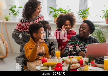 ragazza allegra afroamericana che agita la mano durante la videochiamata vicino alla famiglia Foto Stock