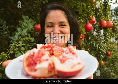 sorridente giovane donna mani che tiene piatto con pezzi freschi di melograno raccolti da albero sullo sfondo con semi rossi. concetto di mangiare autunno fr Foto Stock