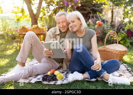 Ritratto di amorevole coppia anziana seduta su coperta durante il picnic e utilizzando tablet digitale, riposo all'aperto Foto Stock