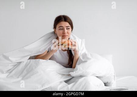 Triste sconvolto depresso donna millenaria europea che mangia hamburger in letto sotto il piumone solo a casa Foto Stock