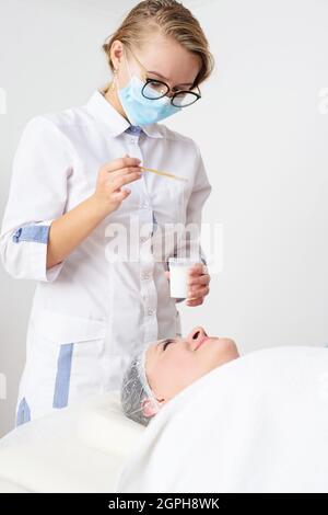 Giovane cosmetologo femminile applica il prodotto cosmetico sul volto della paziente femminile. Centro benessere, procedura cosmetica Foto Stock