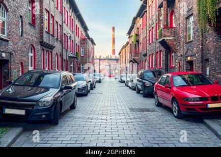 Tradizionale, silesian distretto Nikiszowiec. Ristrutturato, vecchie case in mattoni, strada acciottolata, auto parcheggiate sulla strada, camino in fabbrica nel Foto Stock