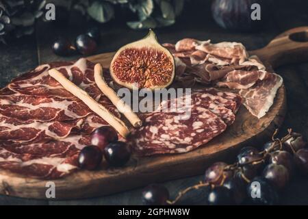 Varie specialità di salsiccia affumicata su un asse di legno, decorate con fichi e uva Foto Stock