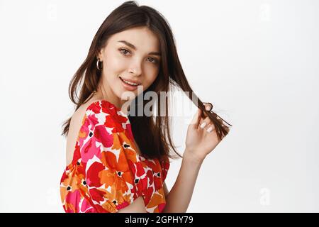 Primo piano ritratto di bella ragazza naturale, giocando con capelli filo, guardando cocquettish e flirty alla macchina fotografica, in posa in abito floreale estate contro Foto Stock