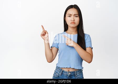 Moody asian ragazza puntando le dita in alto a sinistra angolo, accigliato e accigliato sconvolto da qualcosa di cattivo da parte, in piedi su sfondo bianco Foto Stock