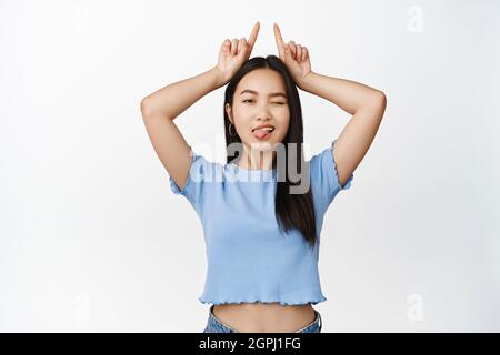 Bella donna asiatica che vana, mostrando corna gesto, puntando in su, in piedi su sfondo bianco Foto Stock