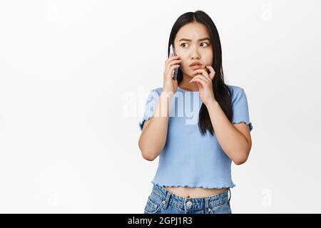 Giovane donna asiatica che guarda disturbata durante la telefonata, ansiosa mentre parla sul cellulare, sfondo bianco Foto Stock