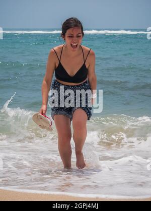 Young Latina Holding un paio di sandali e camminare lungo il Seashore Laughing a Lot Foto Stock