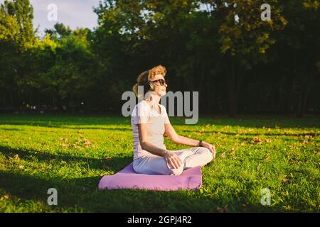 Donna anziana meditando ed esercitando la posizione del loto di yoga all'aperto. Donna Ederly che fa esercizi di stretching su tappeto yoga nel parco sul prato verde al sole Foto Stock