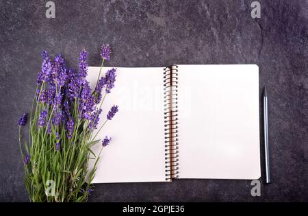 Vista dall'alto del blocco note vuoto aperto con bouquet di lavanda su sfondo di pietra nera, spazio di copia per il testo Foto Stock