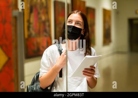 La ragazza messa a fuoco in una maschera protettiva nel museo guarda l'esposizione situata dietro il vetro Foto Stock