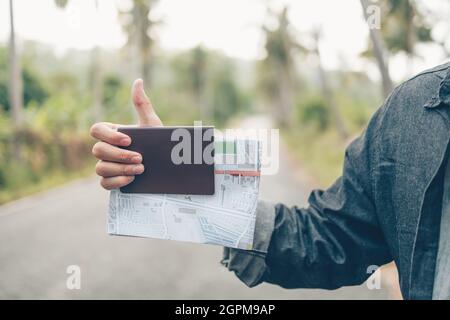 Uomo con zaino tenere la mappa mano segno 'Hitchhiking' e lui a piedi sulla strada nella foresta. Concetto di viaggio zaino. Foto Stock